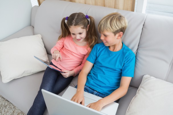 Happy siblings using laptop and tablet pc on sofa at home in the living room