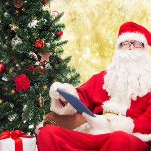 technology, holidays and people concept - man in costume of santa claus with tablet pc computer, gifts and christmas tree sitting in armchair over yellow lights background